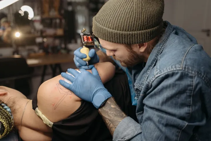 A tattoo artist in a denim jacket working with blue gloves